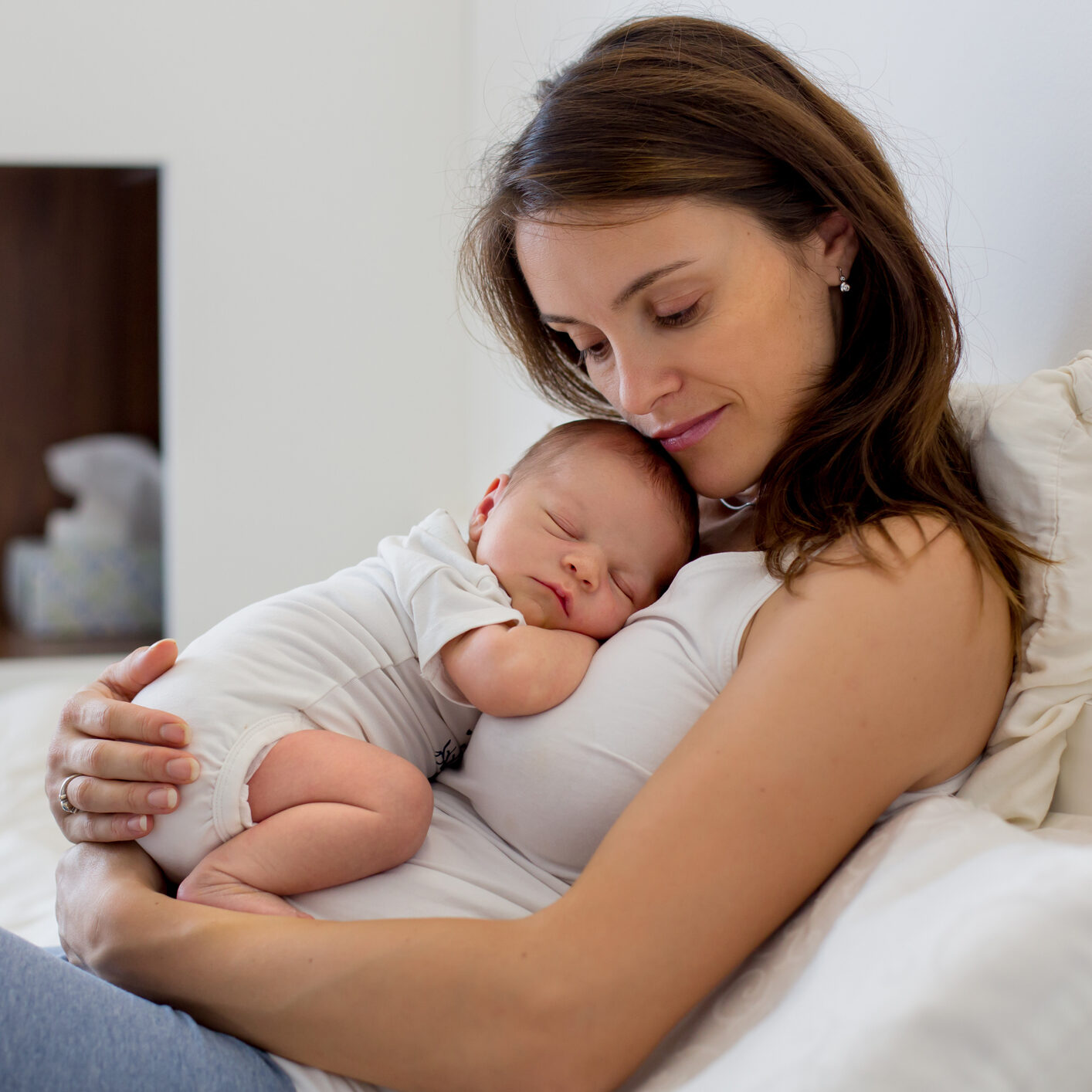 Young mother lying in bed with her newborn baby boy Young mother lying in bed with her newborn baby boy, holding him in her arms and smiling from happiness